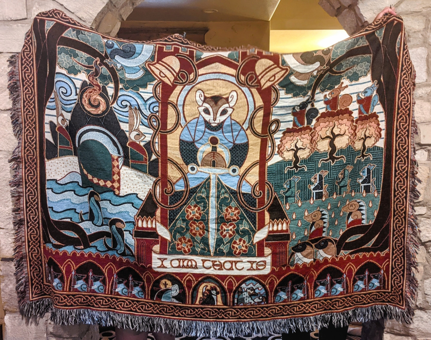 Decorative tapestry with intricate designs and characters, hanging against a stone wall.